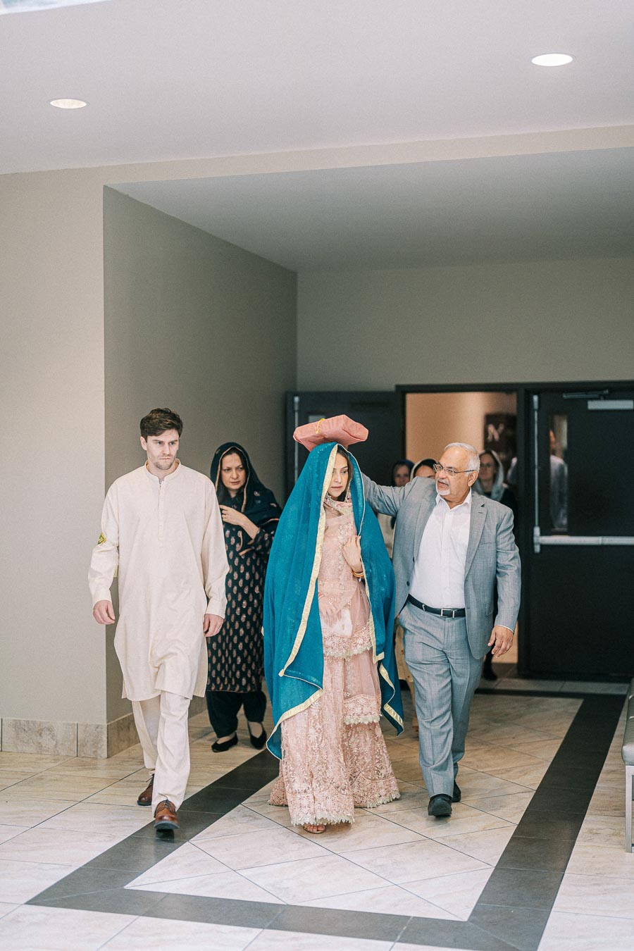 A bride dressed in traditional South Asian attire, including a pink lehenga and blue dupatta, walks through a hallway accompanied by family members, highlighting cultural wedding traditions.