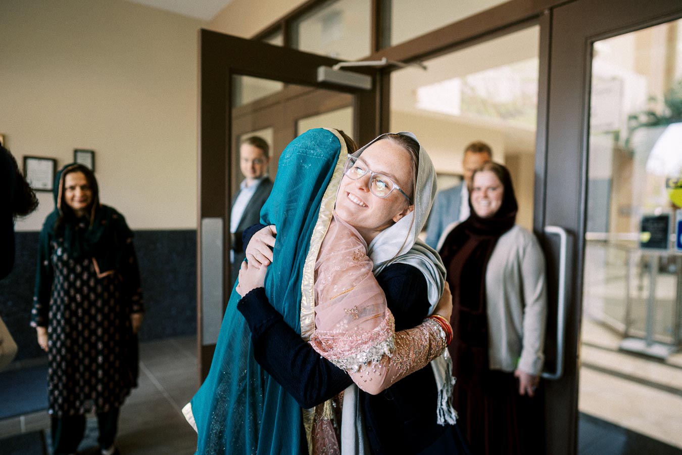 Two women embrace warmly at an indoor gathering, wearing traditional attire with headscarves. Other people stand smiling in the background near a doorway, creating a welcoming and joyful atmosphere.