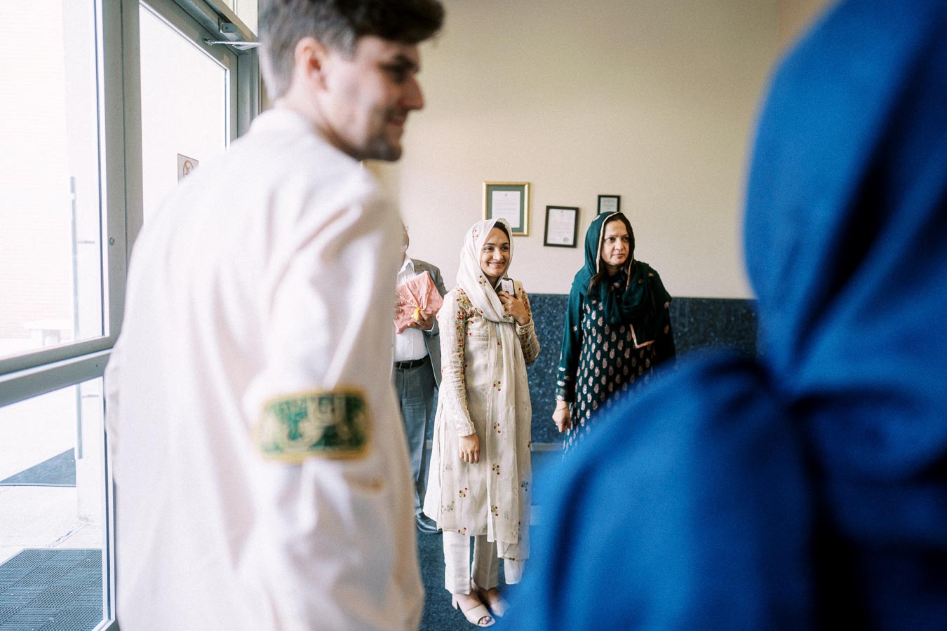 A group of people standing indoors, focused on a cheerful woman in a patterned headscarf and traditional attire, holding a phone. Some are wearing a blue garment. Sunlight enters through large windows, creating a warm atmosphere.