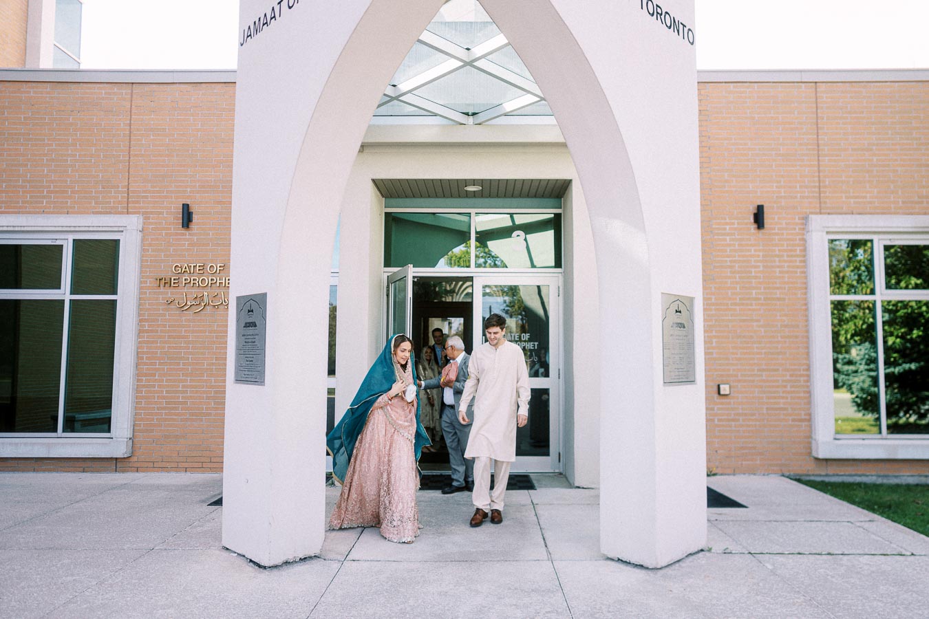 A couple exits a mosque through an arched entrance marked Gate of the Prophet in Toronto. The woman is wearing a traditional pink and teal outfit, while the man is dressed in a beige kurta. The mosque's architecture features brick walls and glass doors, creating a serene atmosphere.