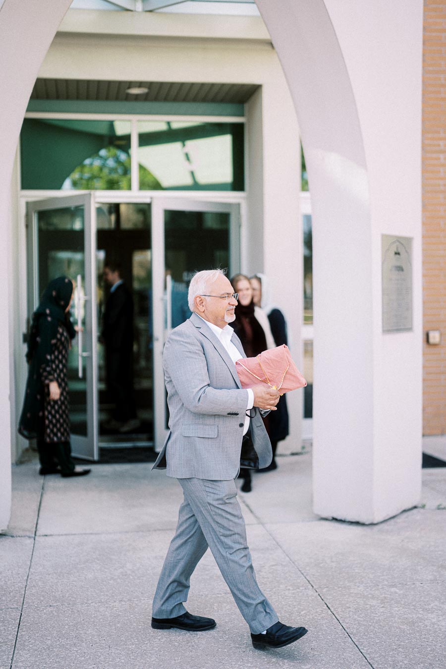 Senior man in a gray suit carrying a package outdoors, with a building entrance and people in the background.