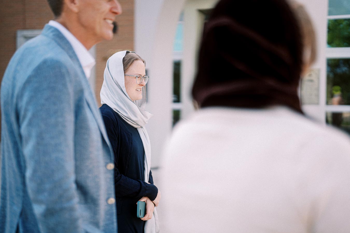 Smiling woman in a light headscarf and glasses holds a phone, standing between two other people in an outdoor setting.