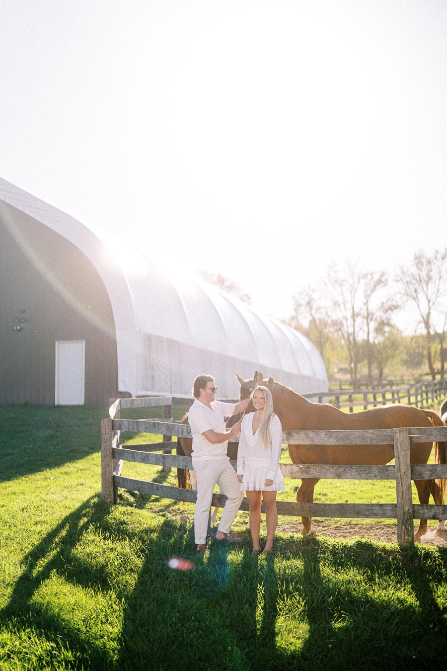 A couple in white outfits standing by a wooden fence with a horse, near a large barn structure, on a sunny day in a grassy field.