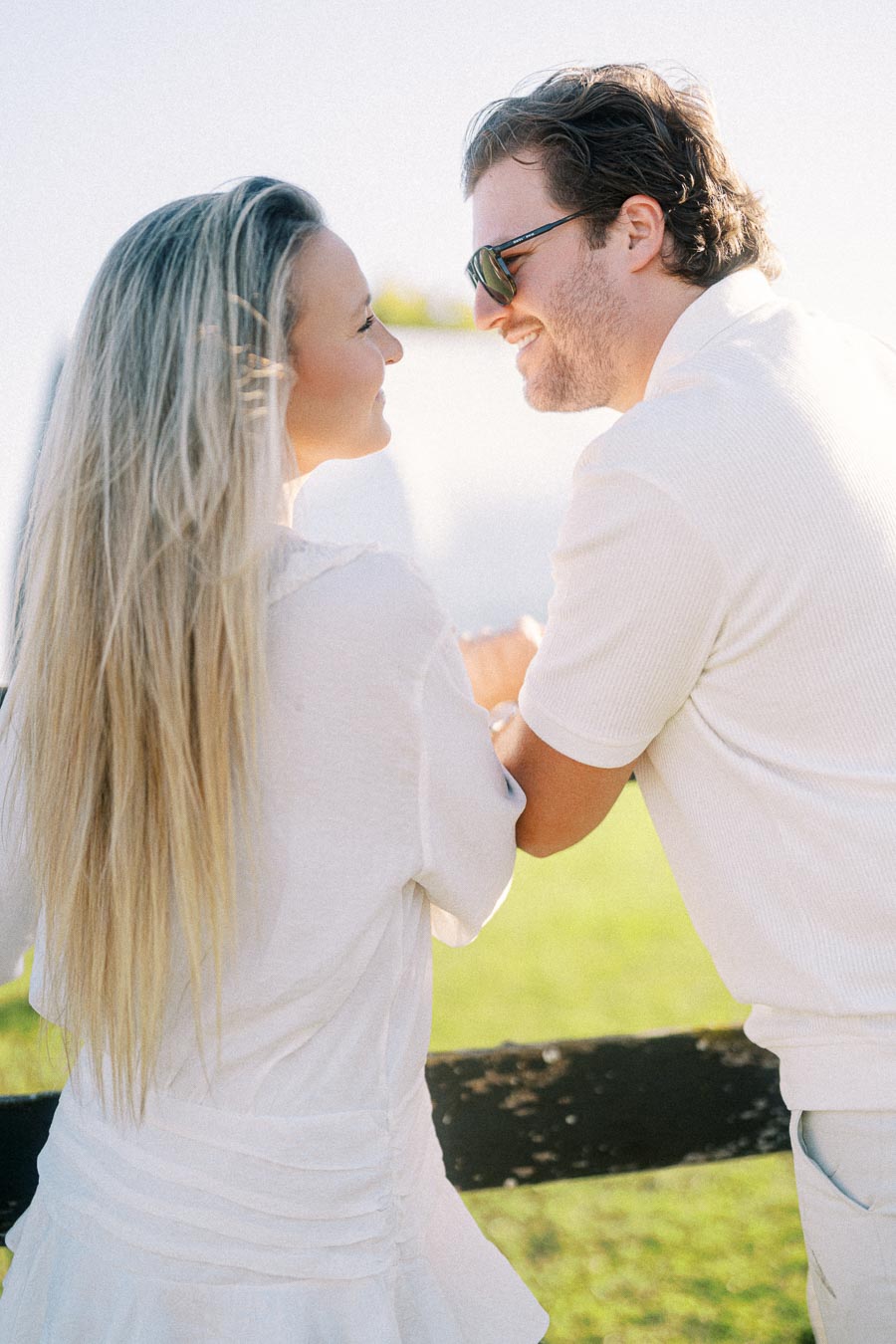 Couple enjoying a sunny day outdoors, smiling at each other while leaning on a fence, wearing casual summer attire in a bright, natural setting.