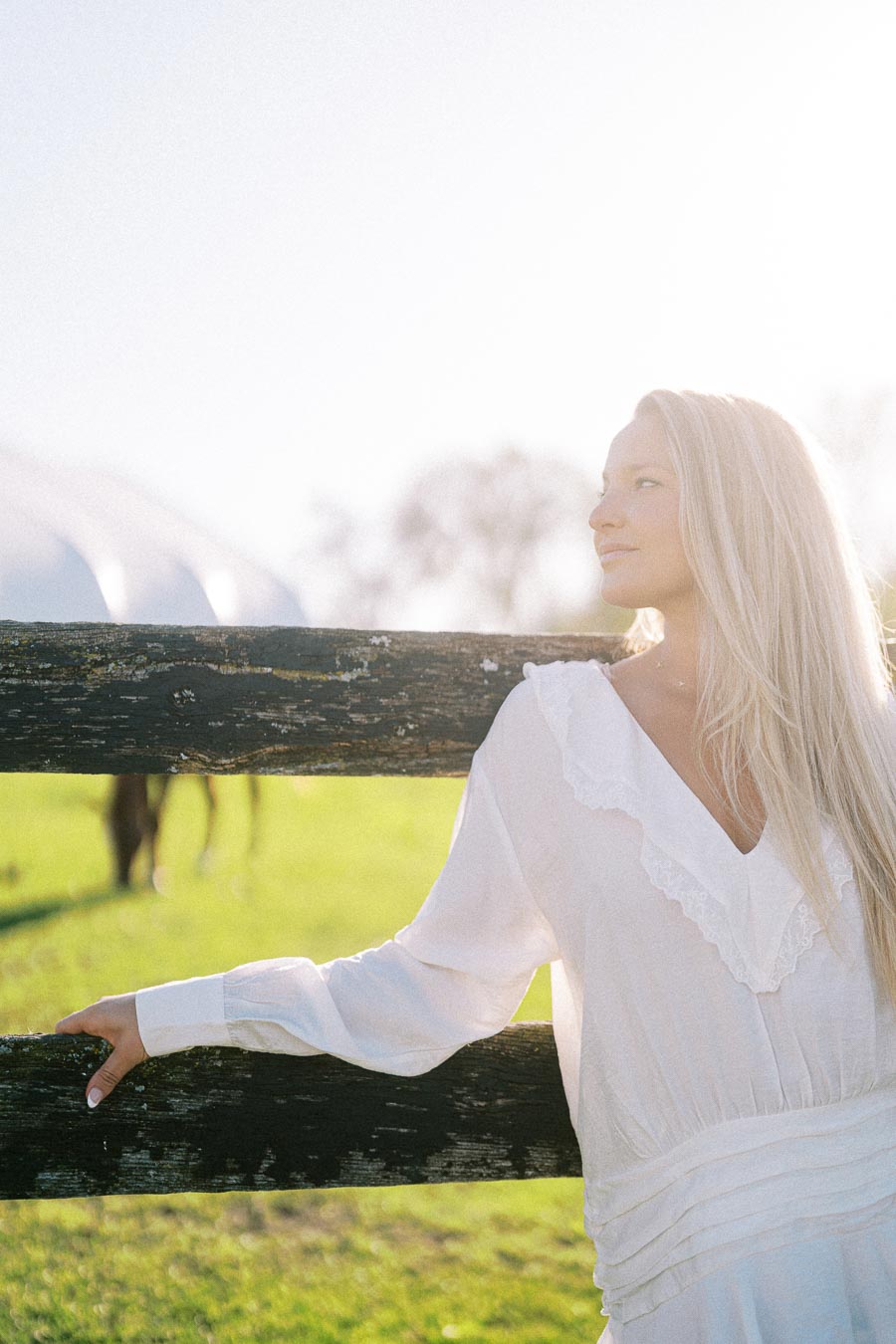 Woman in a white dress standing by a wooden fence in a sunlit field with a blurred horse in the background.