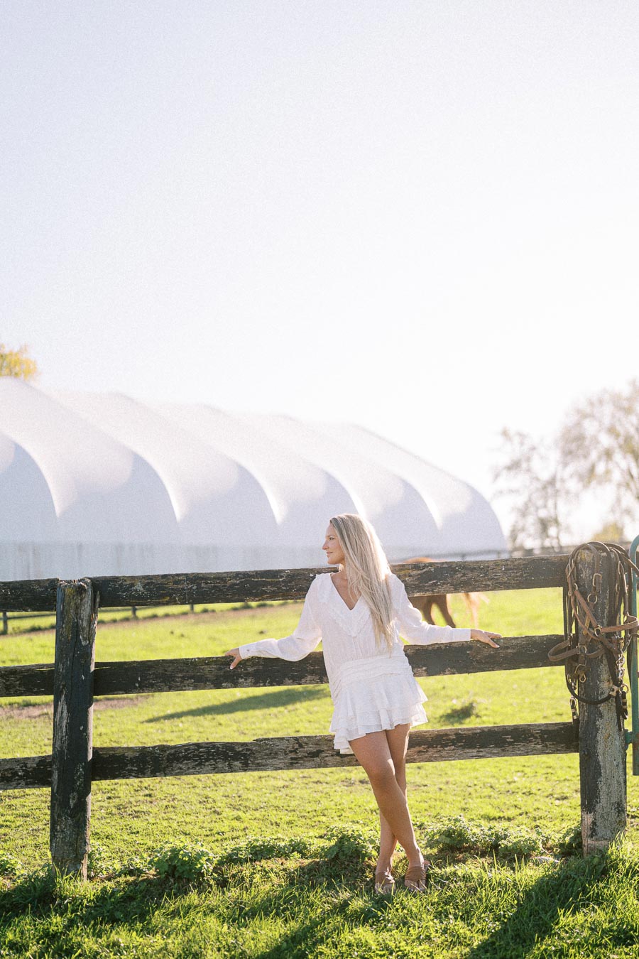 A woman in a white dress standing by a wooden fence in a sunlit field, with a large white building in the background.