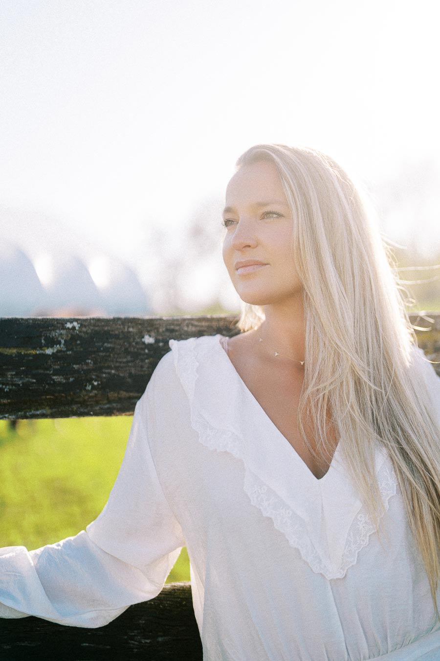 A woman in a white blouse standing against a wooden fence in a sunny rural setting, with long blonde hair and a serene expression.