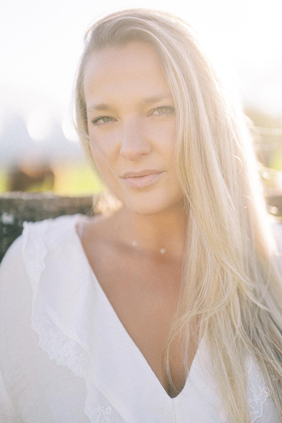 A woman with long blonde hair in a white blouse looking directly at the camera, backlit by sunlight creating a soft glow.
