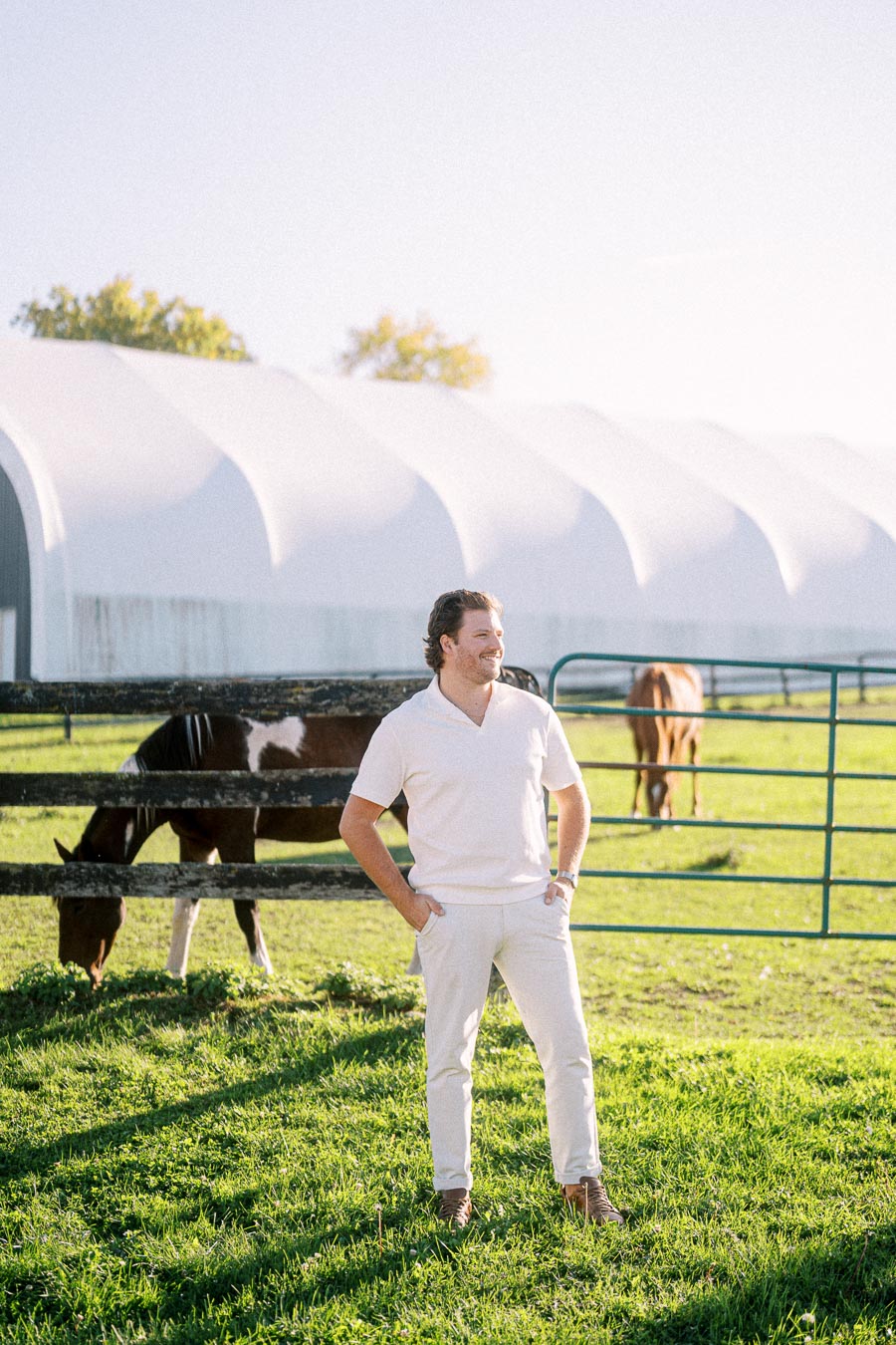 Man in white outfit standing in sunny green pasture with grazing horses, near a white barn structure.