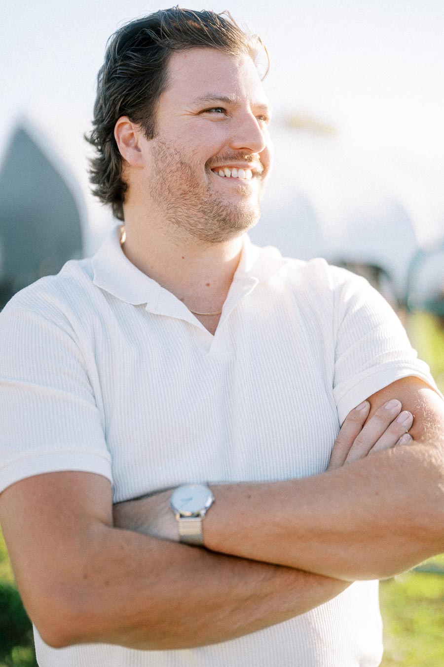 A man smiling outdoors, wearing a white polo shirt and a watch, with arms crossed.