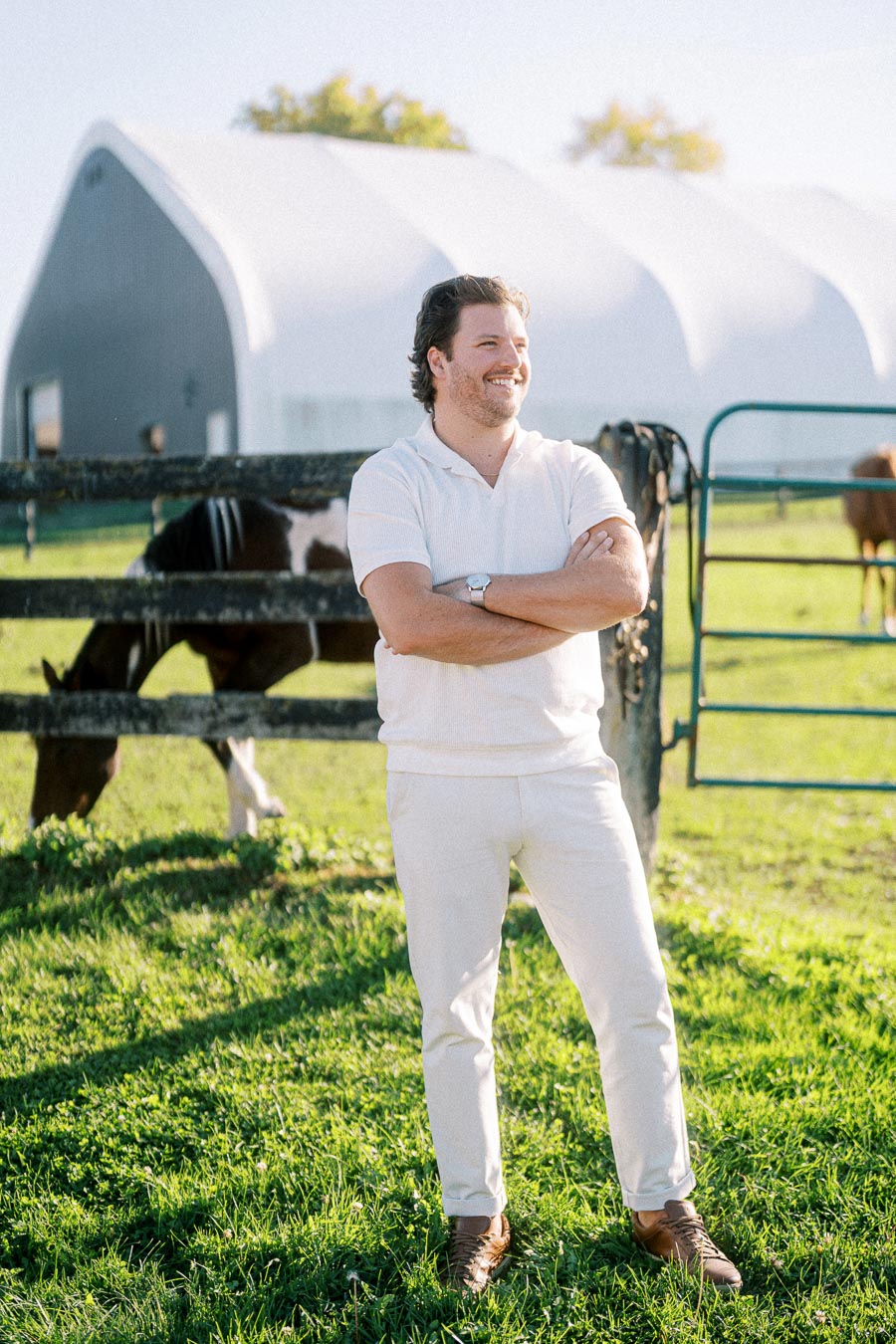 Man in white outfit standing on a sunlit farm with a horse and barn in the background.
