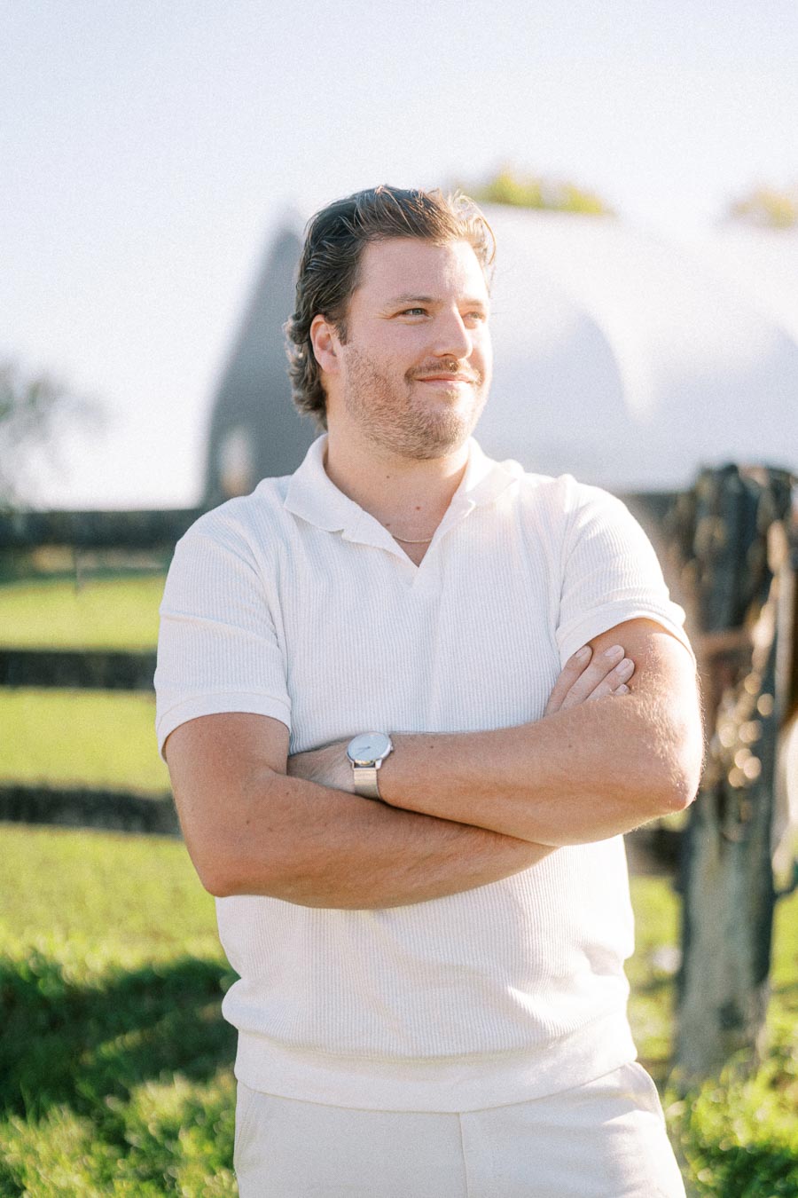Man in a white polo shirt standing outdoors with arms crossed, in a sunny green field.