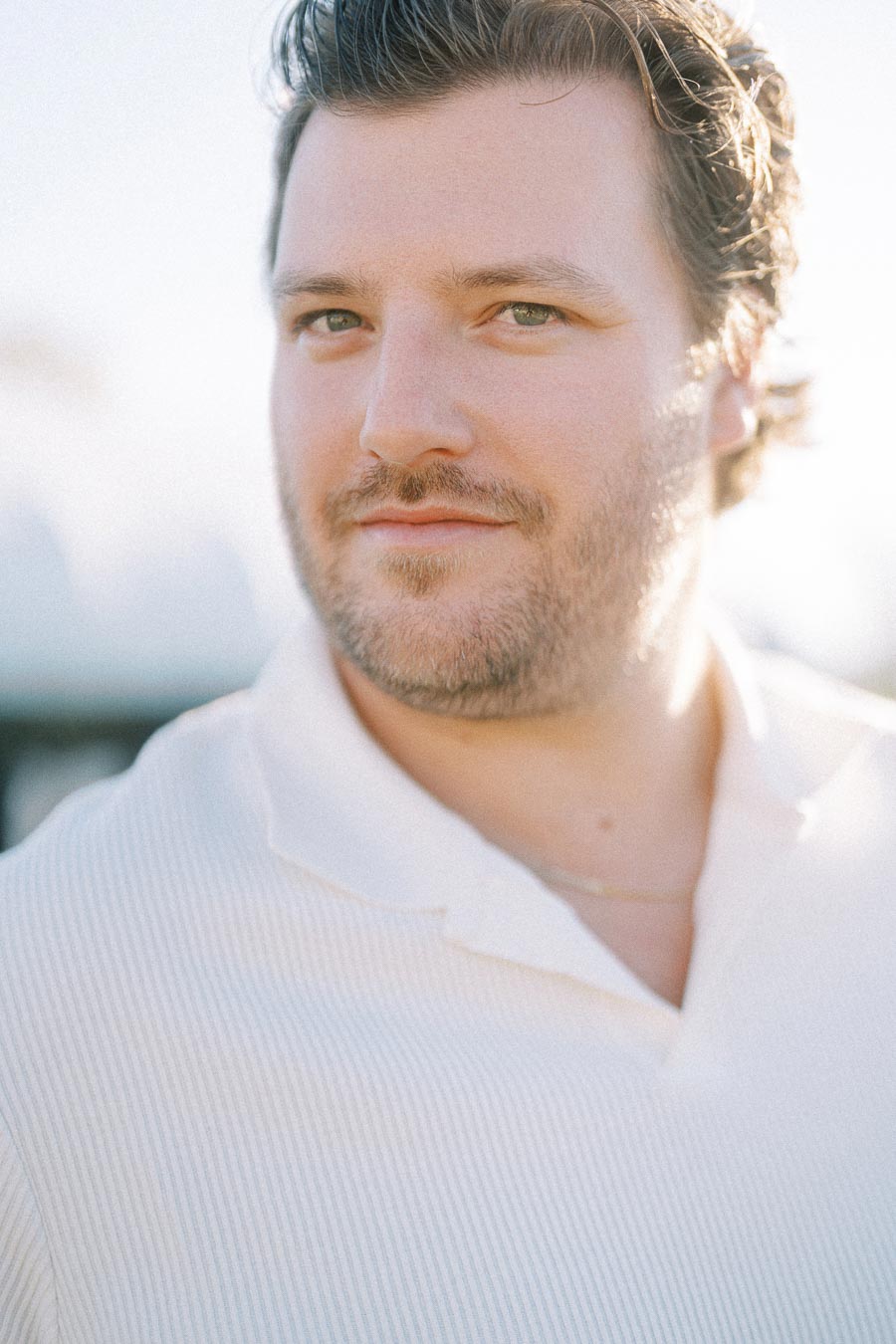 Close-up portrait of a man with short dark hair and a beard, wearing a white shirt and looking slightly to the right, with a blurred outdoor background