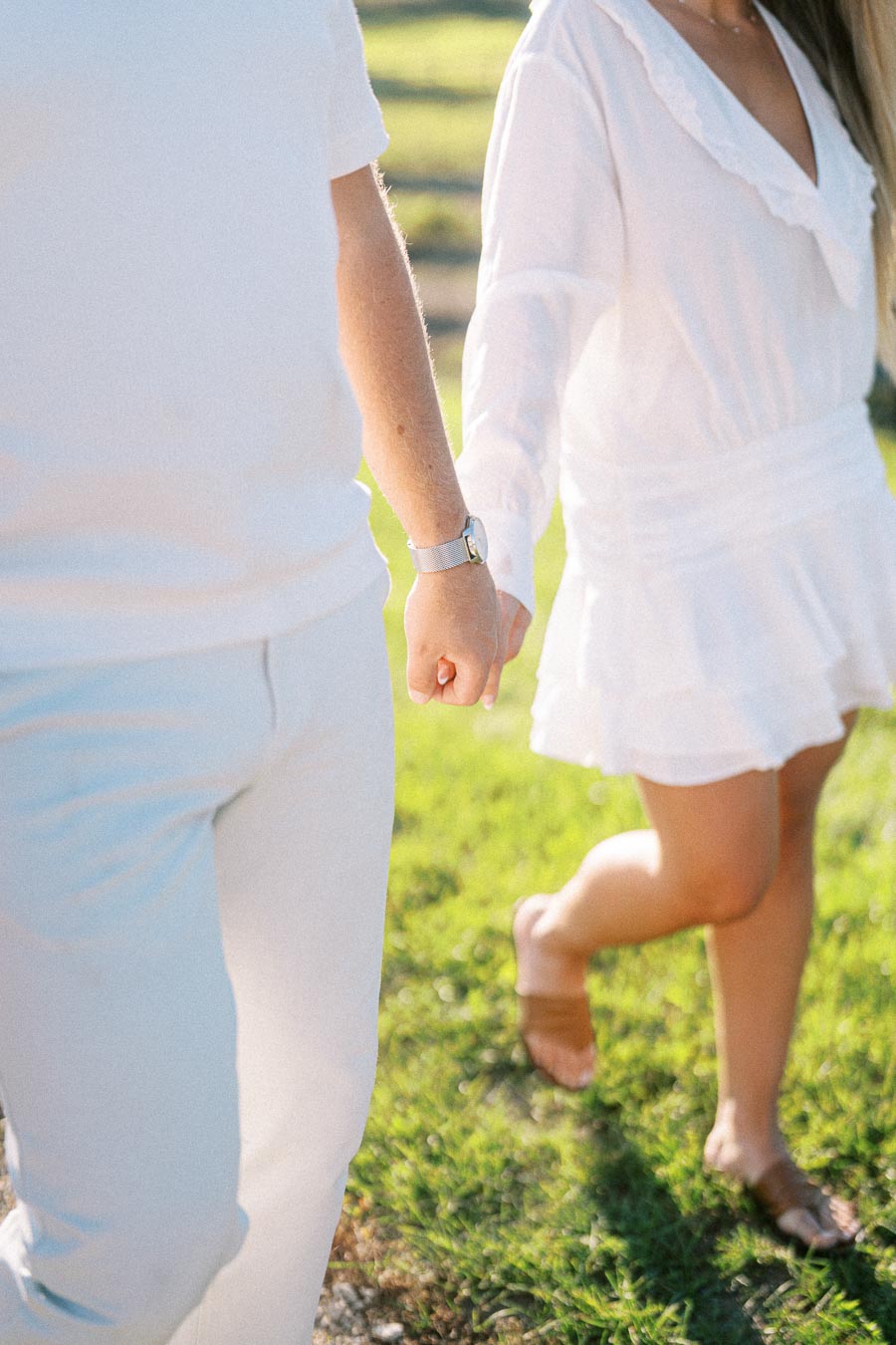 A couple walking hand in hand on a sunny day, wearing white clothing, with lush green grass in the background.