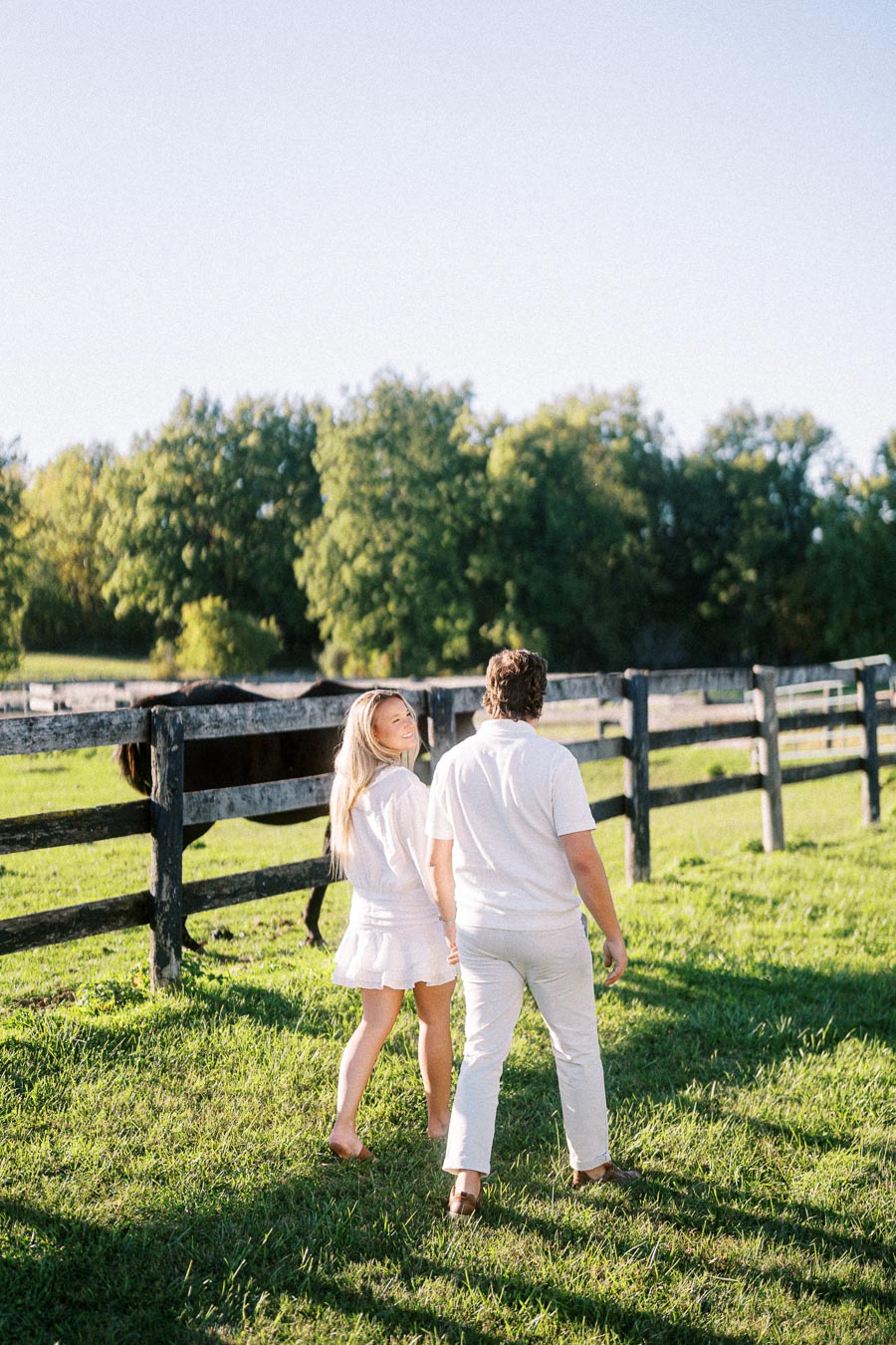A couple dressed in white walking on a grassy field near a wooden fence with a horse, surrounded by lush green trees under a clear blue sky.