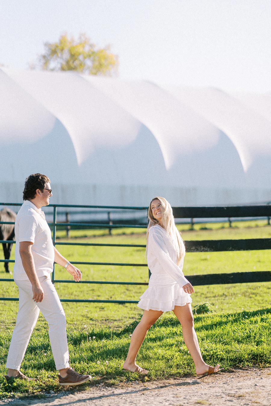 A man and woman dressed in white walk together outside on a sunny day, with a horse and a large white tent visible in the background, creating a serene and joyful atmosphere.