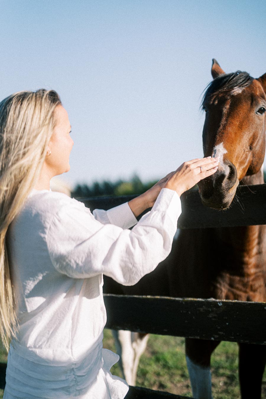 A woman with long blond hair, wearing a white shirt, gently pets a brown horse over a wooden fence in a sunny outdoor setting.