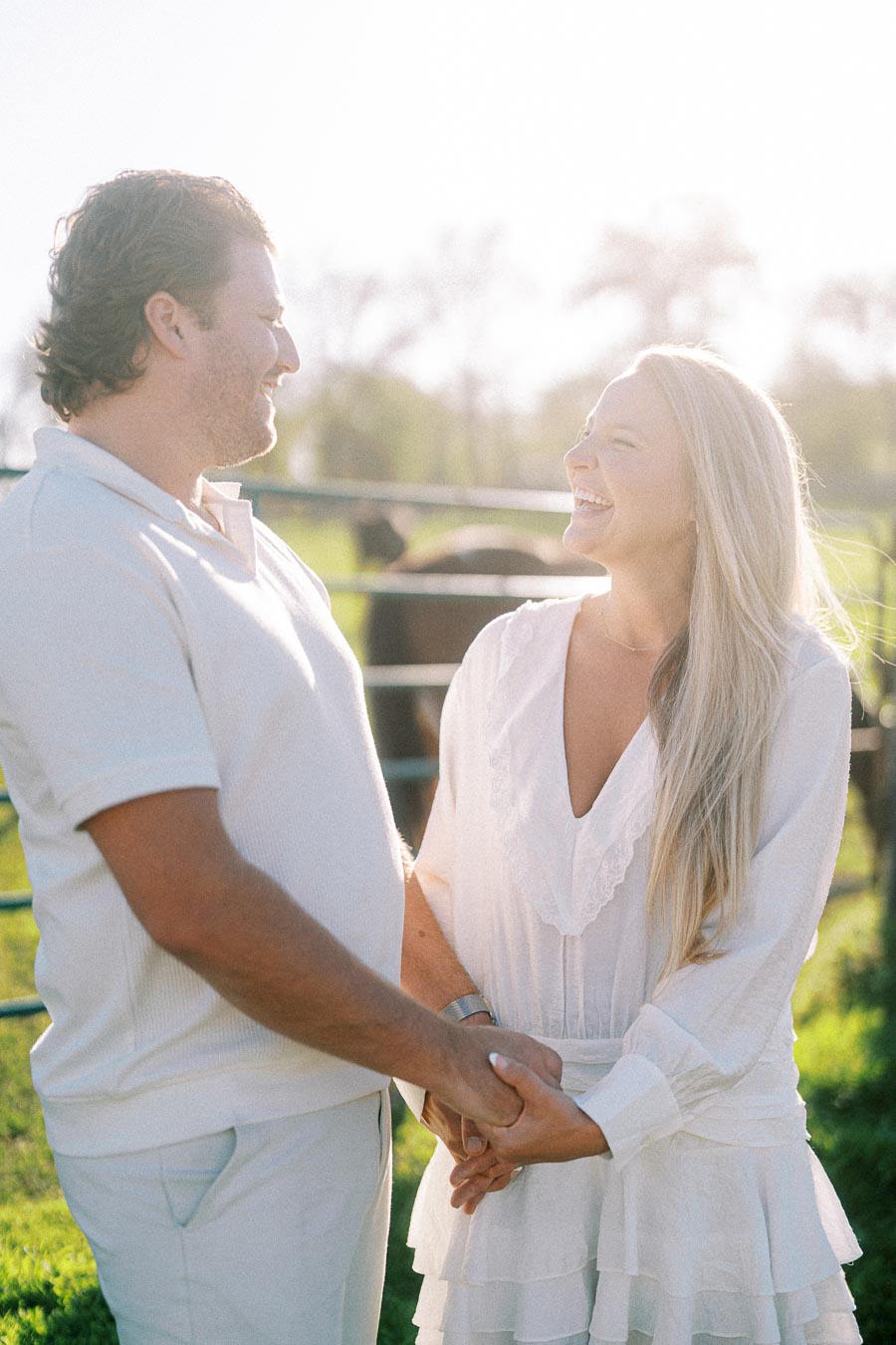 A happy couple in white clothing holding hands and smiling at each other in a sunlit field with a blurred background and fence.