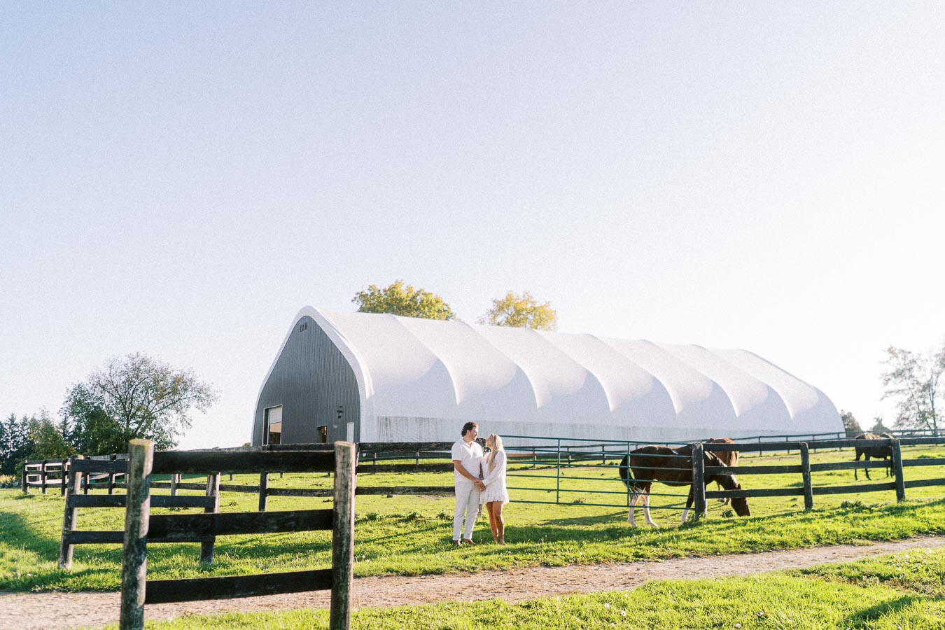 A couple standing in front of a large white barn on a sunny day with grazing horses in the pasture, surrounded by green grass and wooden fences.