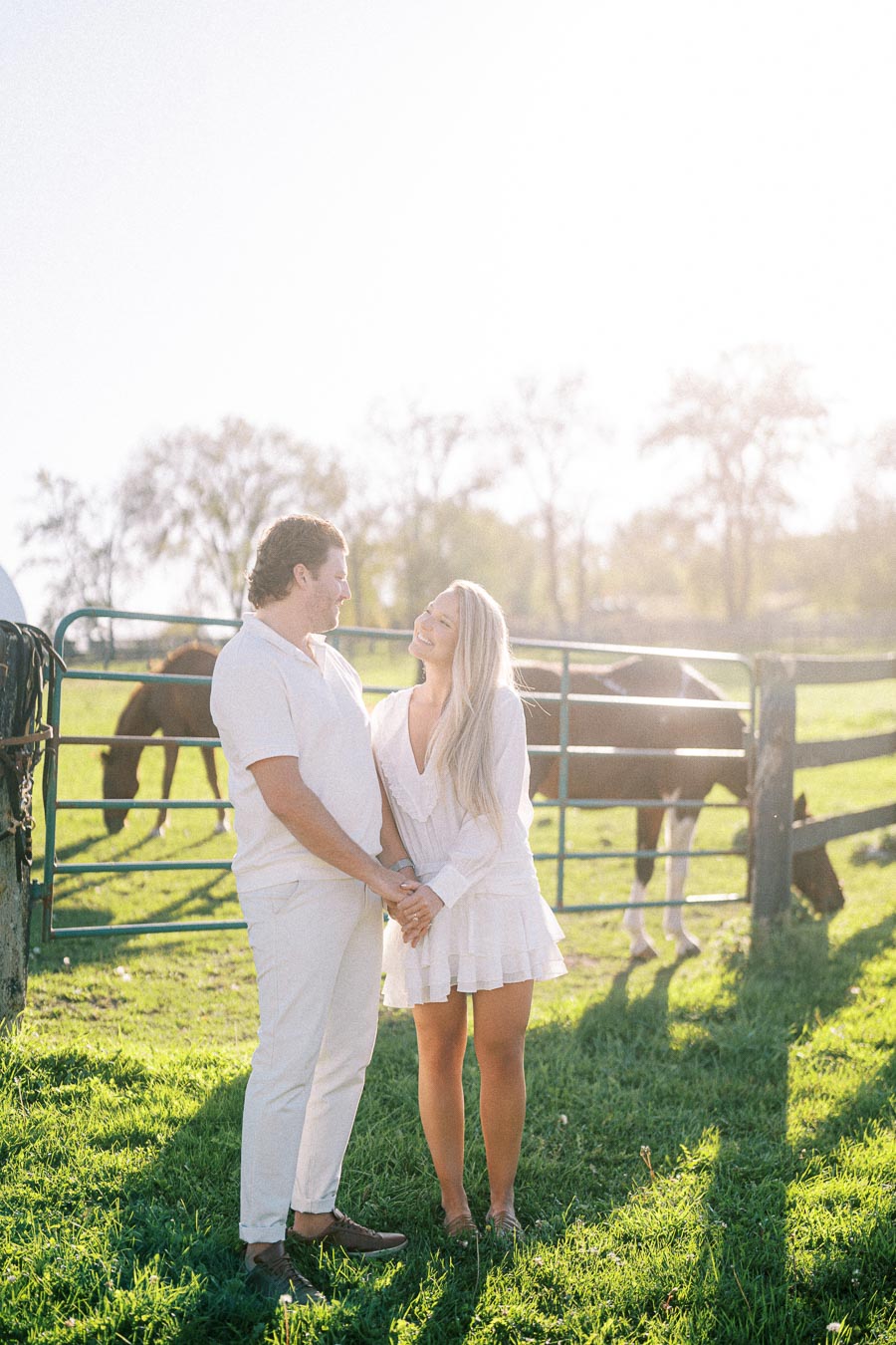 A couple in white attire enjoying a sunny day at a farm, standing in front of a fence with grazing horses and lush greenery in the background.
