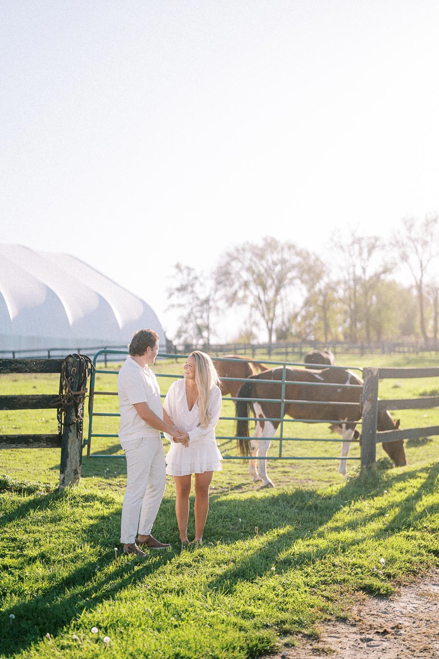 A couple dressed in white standing and holding hands in a sunny pasture with horses behind a fence, surrounded by lush green grass and a bright, clear sky.