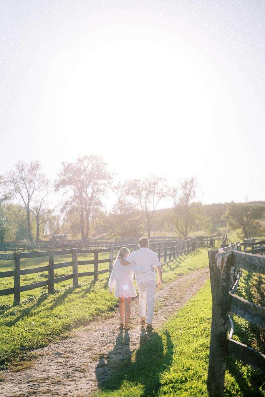 A couple walking arm-in-arm down a sunny rural path, surrounded by a green field and rustic wooden fences, under a bright sky.