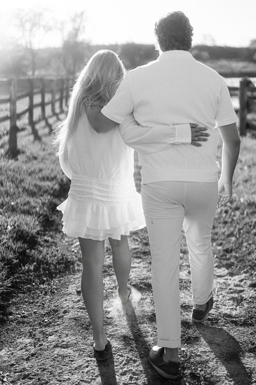A couple walking arm in arm down a rustic dirt path lined with wooden fences, captured in a black and white photograph. The scene conveys a sense of romance and serenity in a countryside setting.