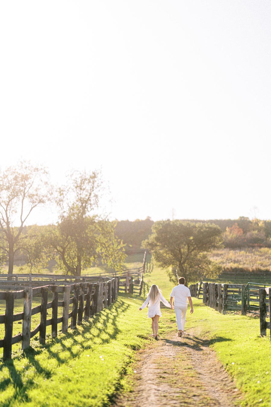 A couple holding hands and walking down a sunlit dirt path in a scenic countryside setting, surrounded by wooden fences and lush greenery.