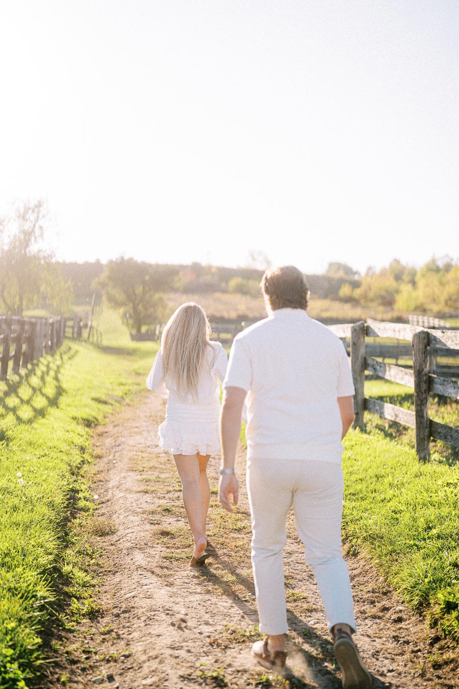 Couple walking along a sunny countryside path bordered by wooden fences, dressed in white, surrounded by lush green grass and illuminated by soft sunlight.
