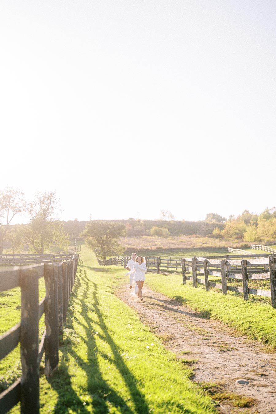 Two people running down a sunlit dirt path in a picturesque rural landscape, surrounded by wooden fences and lush greenery.