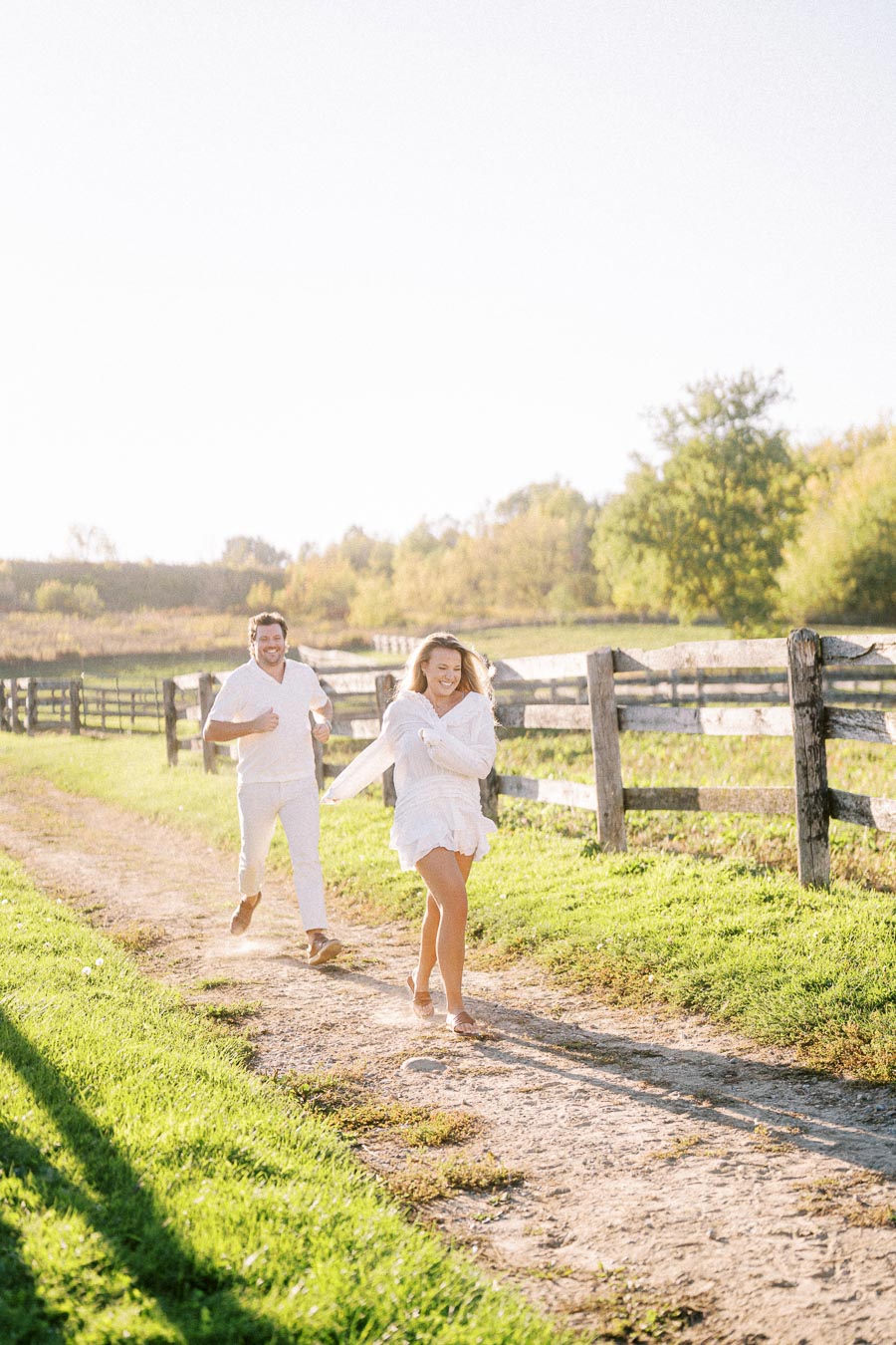 A couple joyfully running on a sunlit dirt path along a wooden fence, surrounded by lush greenery and a clear blue sky.