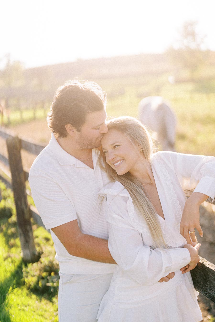 A happy couple enjoying a sunny day in the countryside, standing by a wooden fence with green fields in the background.