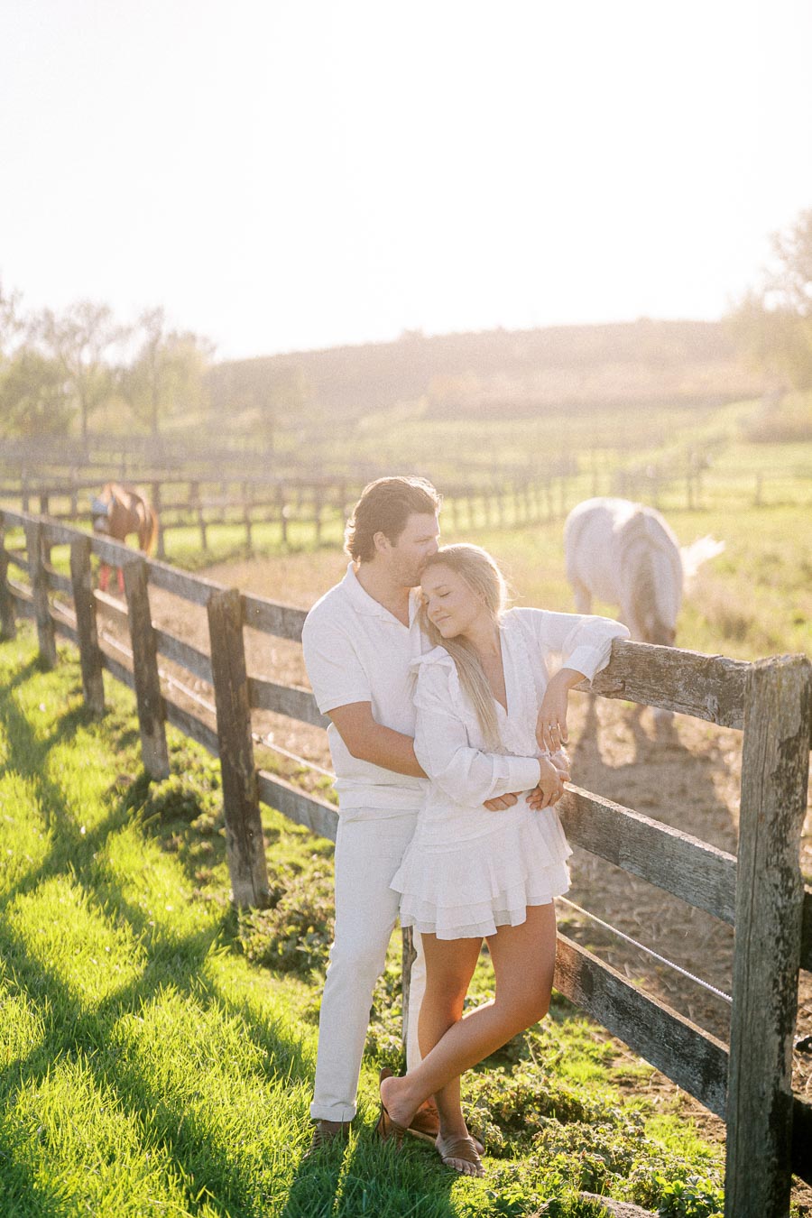 A couple in white clothing embrace lovingly by a rustic wooden fence in a sunny countryside setting, with horses grazing in the background, showcasing a romantic outdoor scene.