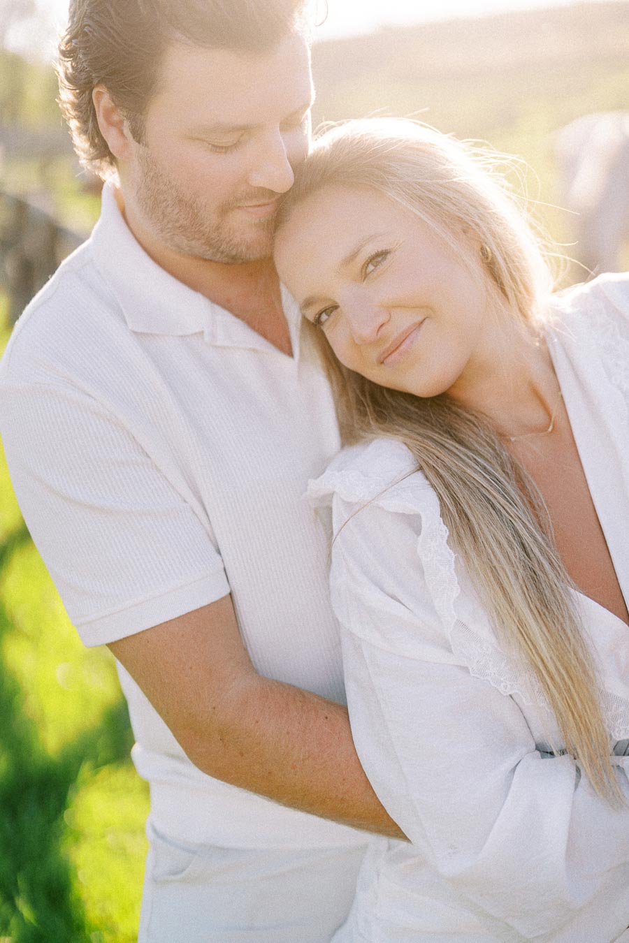 A couple embracing in a sunlit outdoor setting, wearing white clothing, with the woman smiling and leaning back against the man.