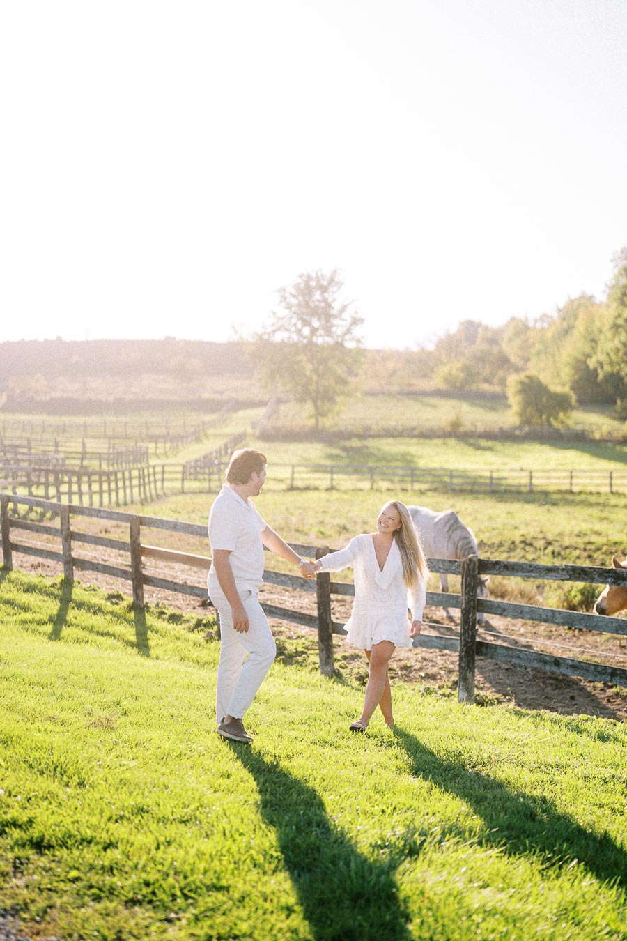 Couple in white outfits walking hand in hand along a scenic, sunlit country pasture with a wooden fence and lush green grass, evoking romance and serenity in a rural setting.