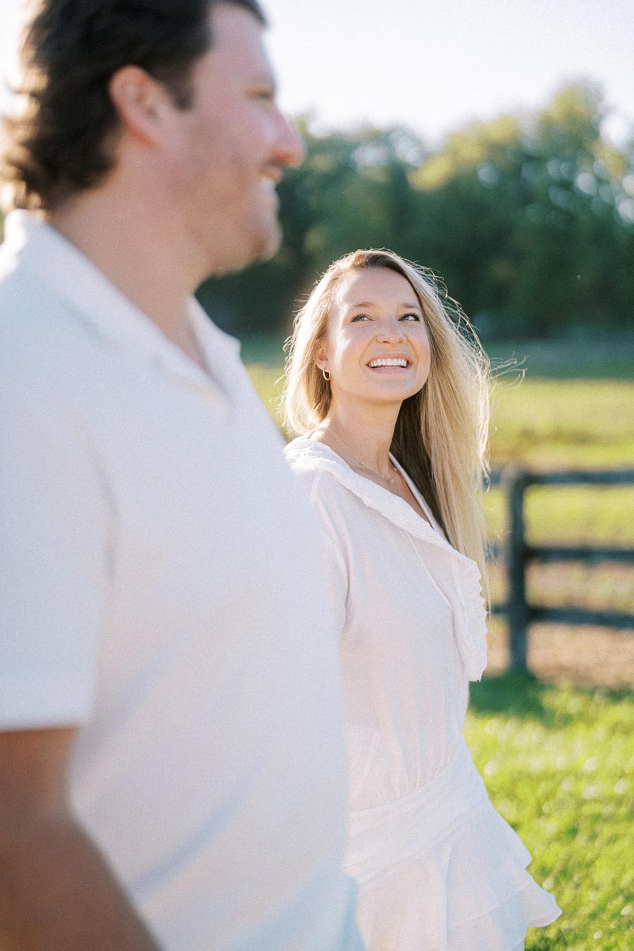 Smiling woman in a white dress walking outdoors with a man, enjoying a sunny day in a lush green park.