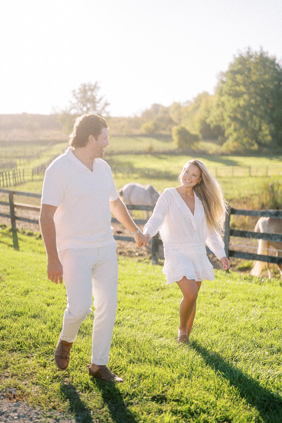 Couple in white outfits walking through a sunlit field next to horses and a wooden fence, enjoying a sunny day outdoors.