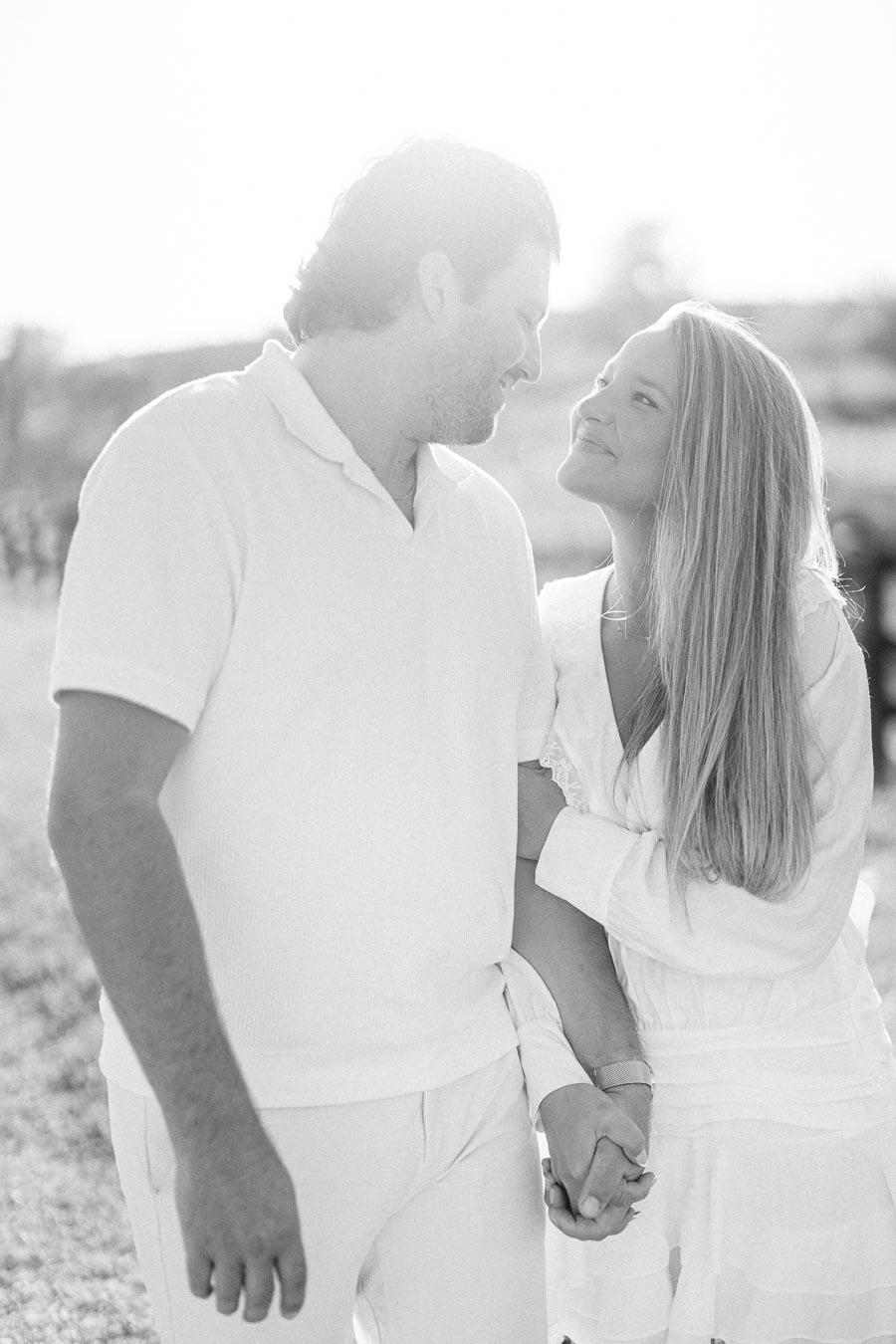 Romantic black and white photo of a couple holding hands and smiling at each other in a sunlit outdoor setting.