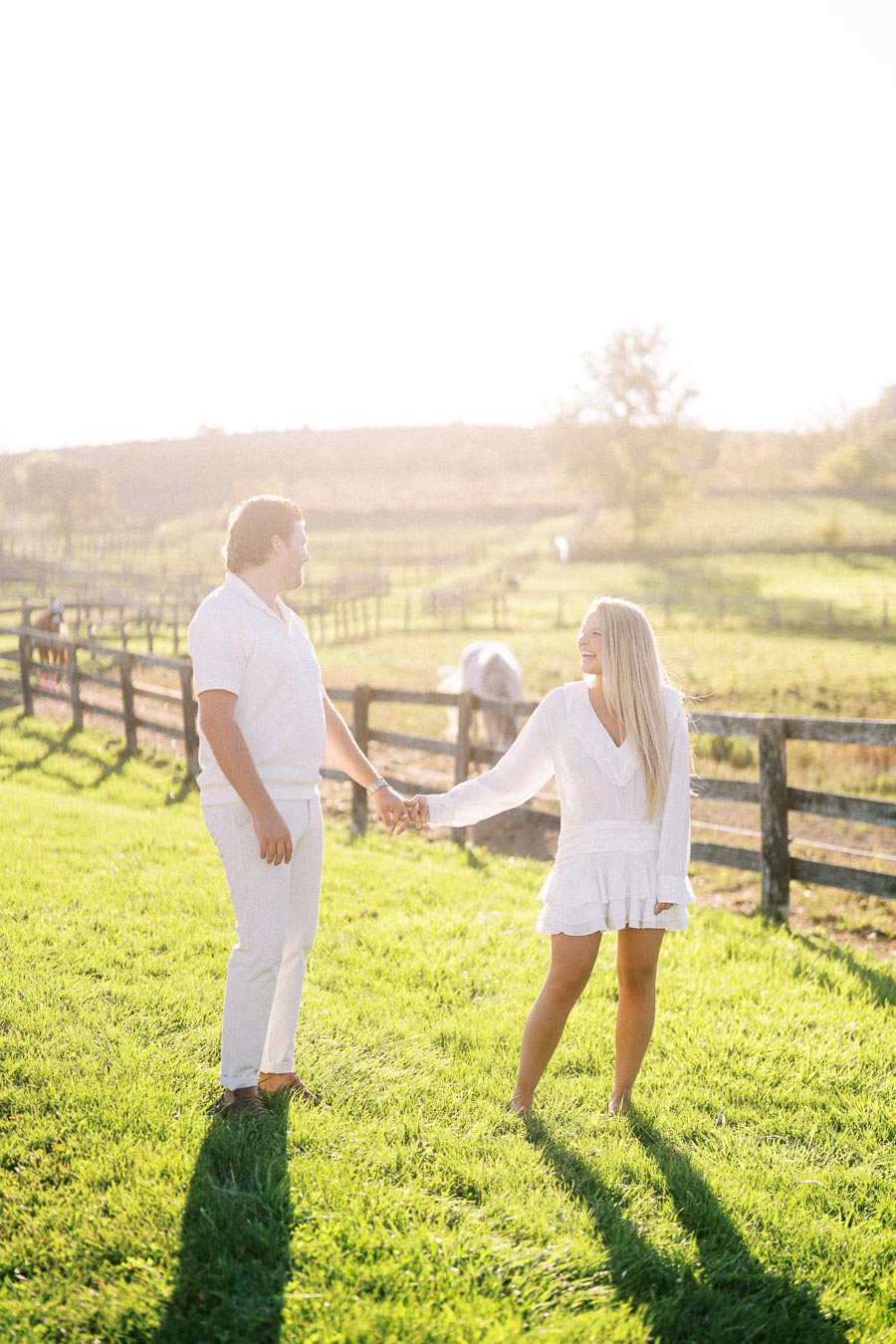 A couple holds hands and smiles in a sunny field with wooden fences in the background, during a bright day, wearing white outfits, illustrating a romantic and serene outdoor moment.