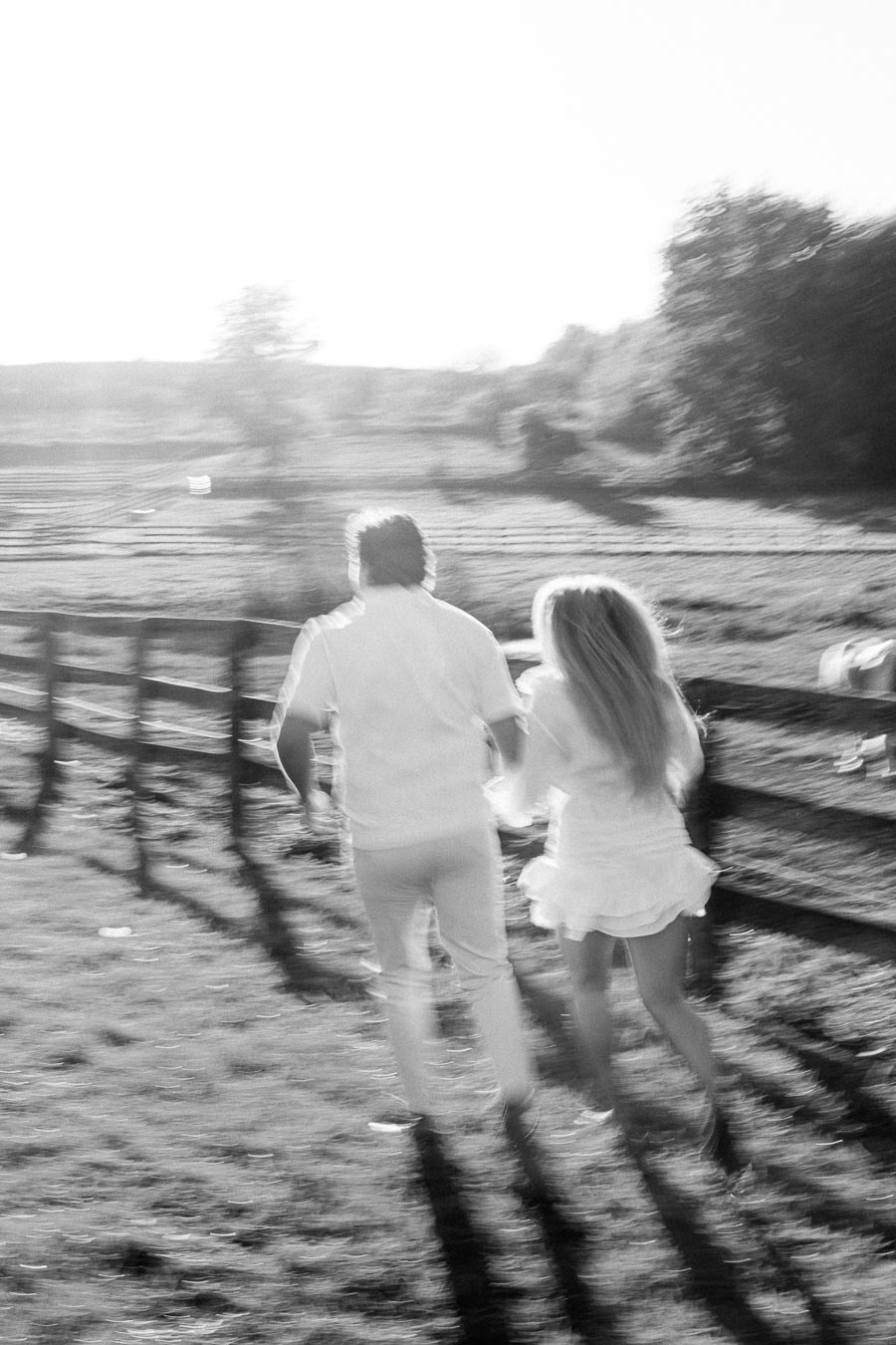A black and white image of a couple running hand in hand through a sunlit field, with a wooden fence and trees in the background, capturing a sense of freedom and joy.