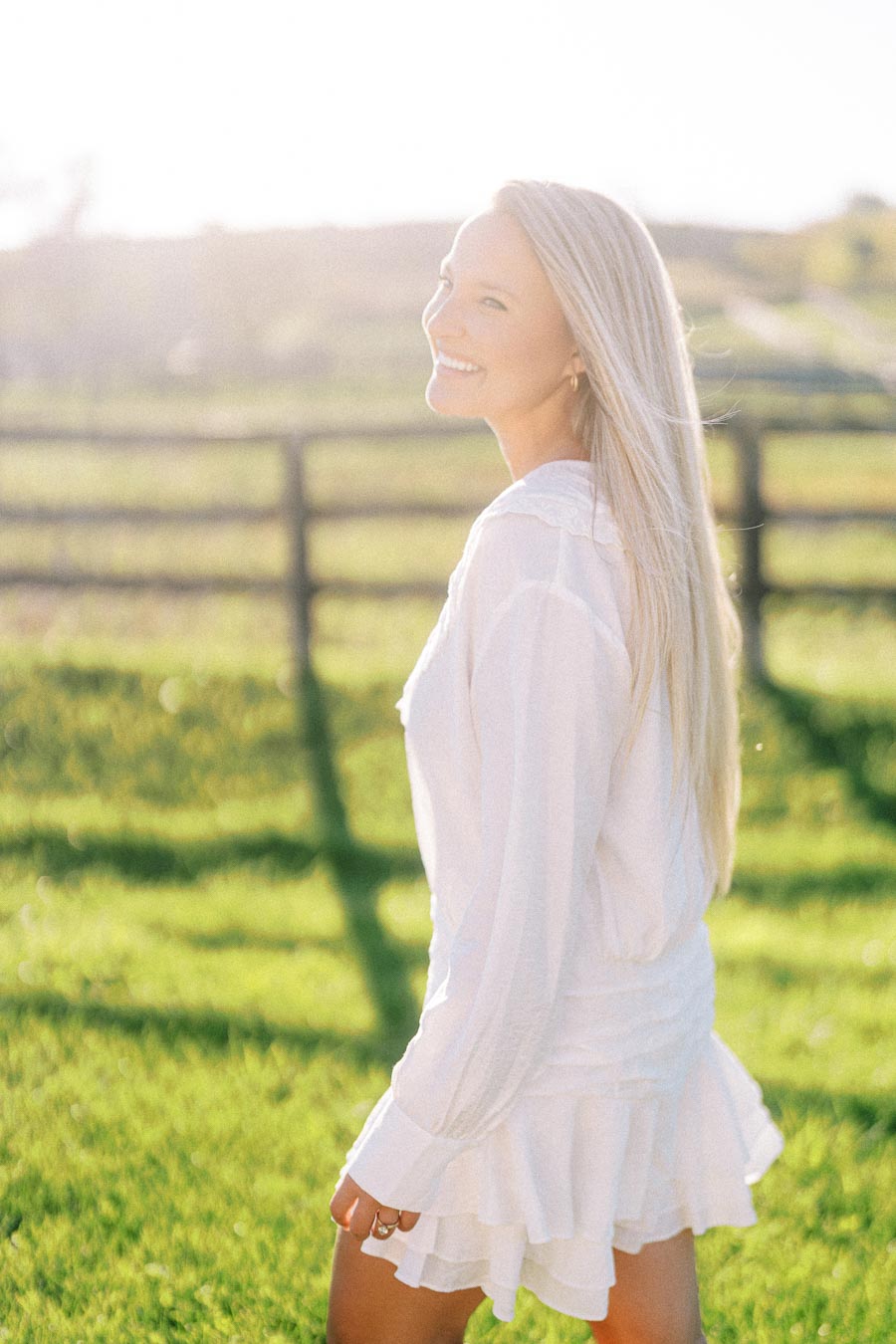 A woman in a white dress smiling and walking in a sunlit green field, with a wooden fence in the background.