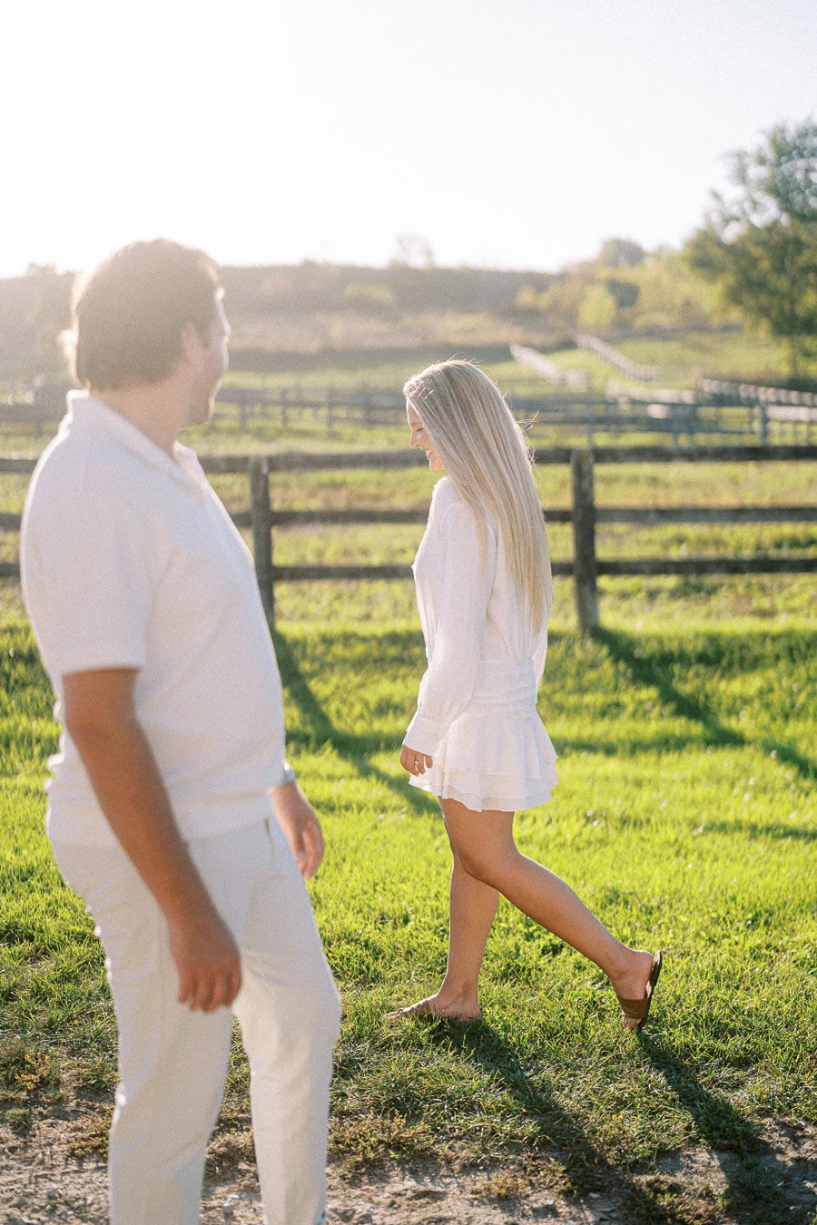 Couple enjoying a sunny day walking in a lush green field with a wooden fence in the background.