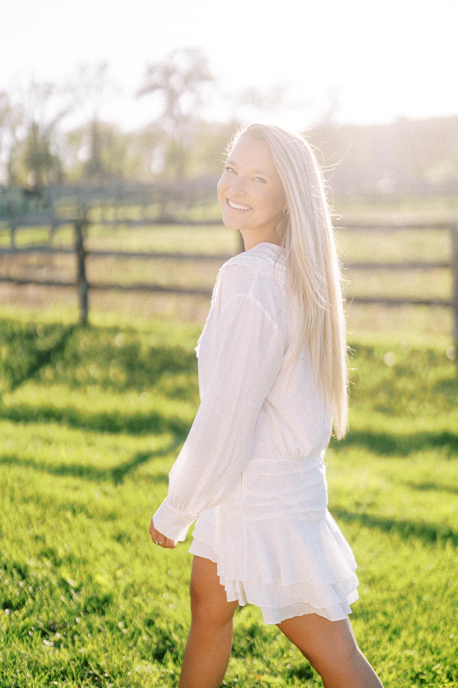 A woman in a white dress smiling and walking in a sunlit field with a wooden fence and trees in the background.