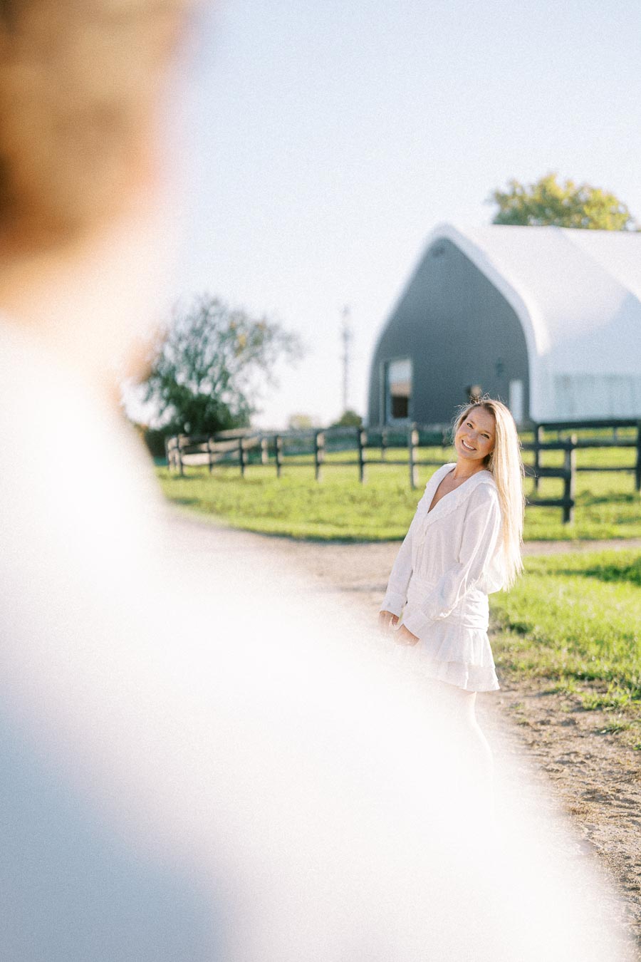 Young woman smiling on a sunny day, standing near a rustic barn on a farm, with green grass and wooden fence in the background.