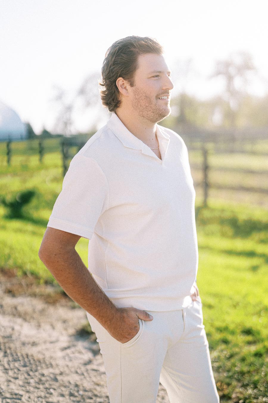 A man in a white shirt standing outdoors in a sunny rural setting, with green grass and a fence in the background.