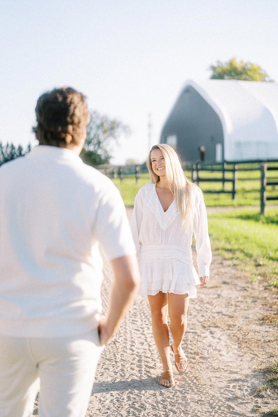 A woman in a white dress smiles while walking towards a man on a sunny day at a rural farm with a white barn in the background.