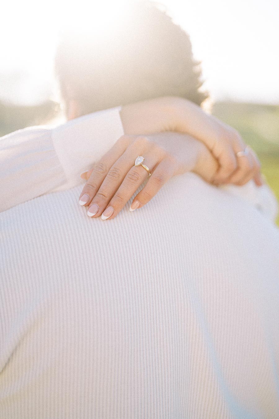 A woman embraces someone in a soft, sunlit setting, displaying a pear-shaped diamond engagement ring on her hand.