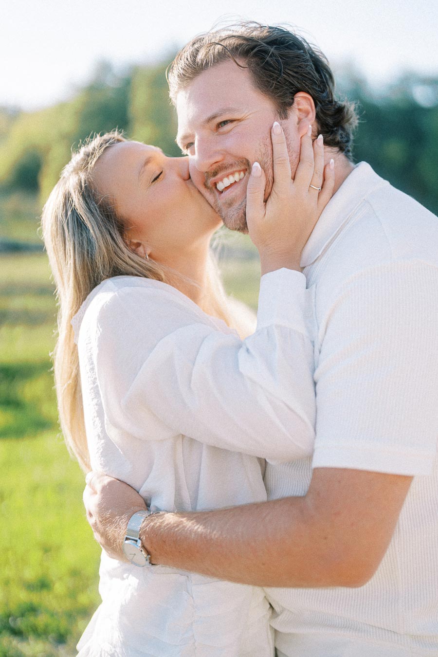 A woman in a white top affectionately kisses a smiling man on the cheek while embracing him in a sunny outdoor setting.