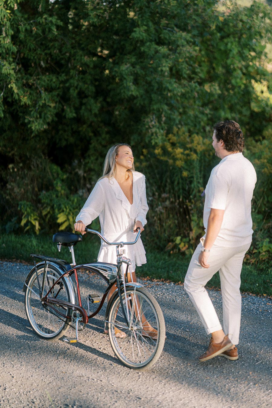A woman smiling while holding a bicycle talks to a man in white clothing on a sunlit path with green trees in the background.