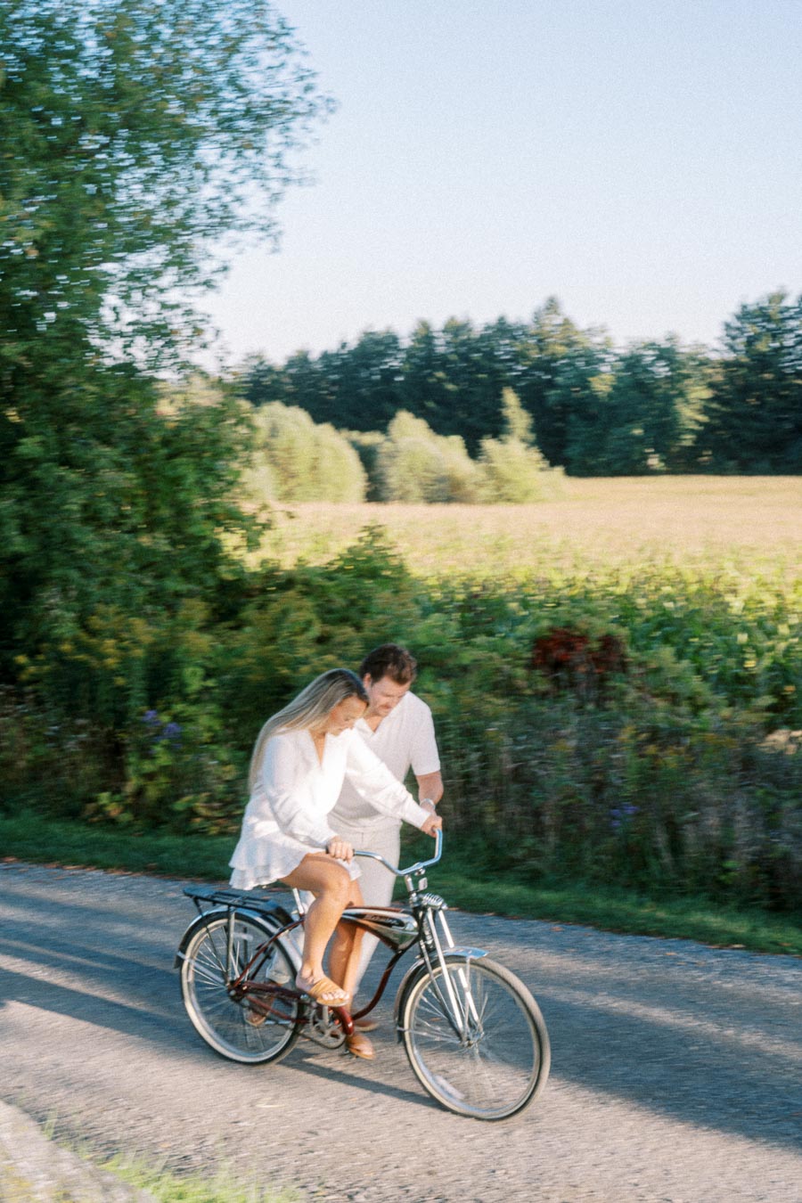 A couple enjoying a leisurely ride on a bicycle down a sunlit rural path, surrounded by lush greenery and open fields.